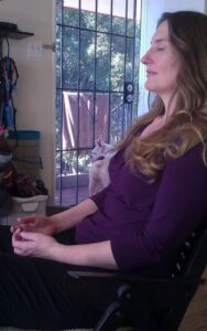 Woman named Sindi sitting in a chair with her eyes closed while meditating. A cute white dog is in the background looking out a screen door on a sunny day in San Diego.